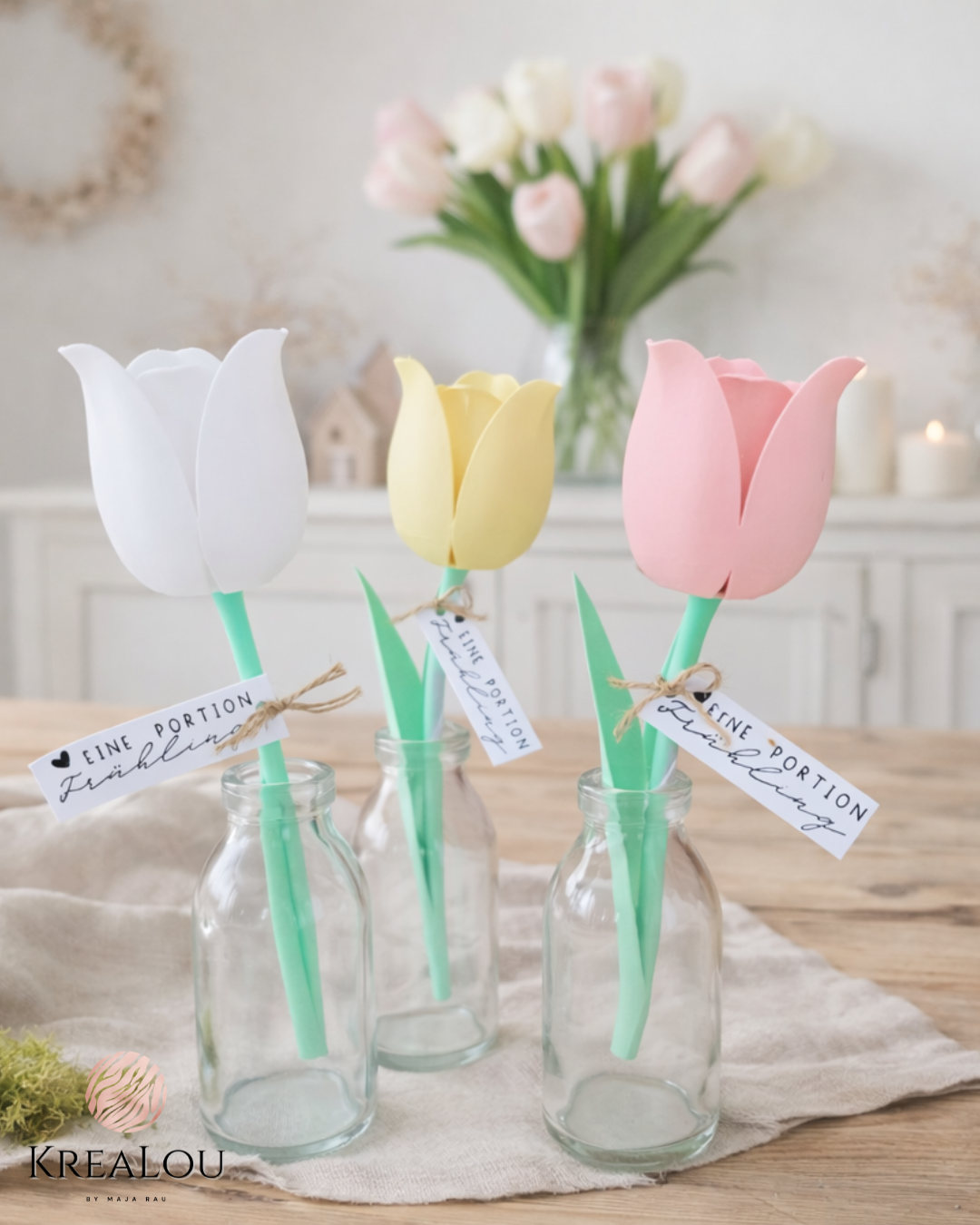 Deko-Tulpen aus PLA in Glasflaschen, mit weißem, gelbem und rosafarbenem Blütenkopf, grünem Stiel und Anhänger „Eine Portion Frühling“, auf hellem Holztisch dekoriert.